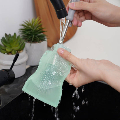 Person washing a green silicone pouch under running water from a faucet.