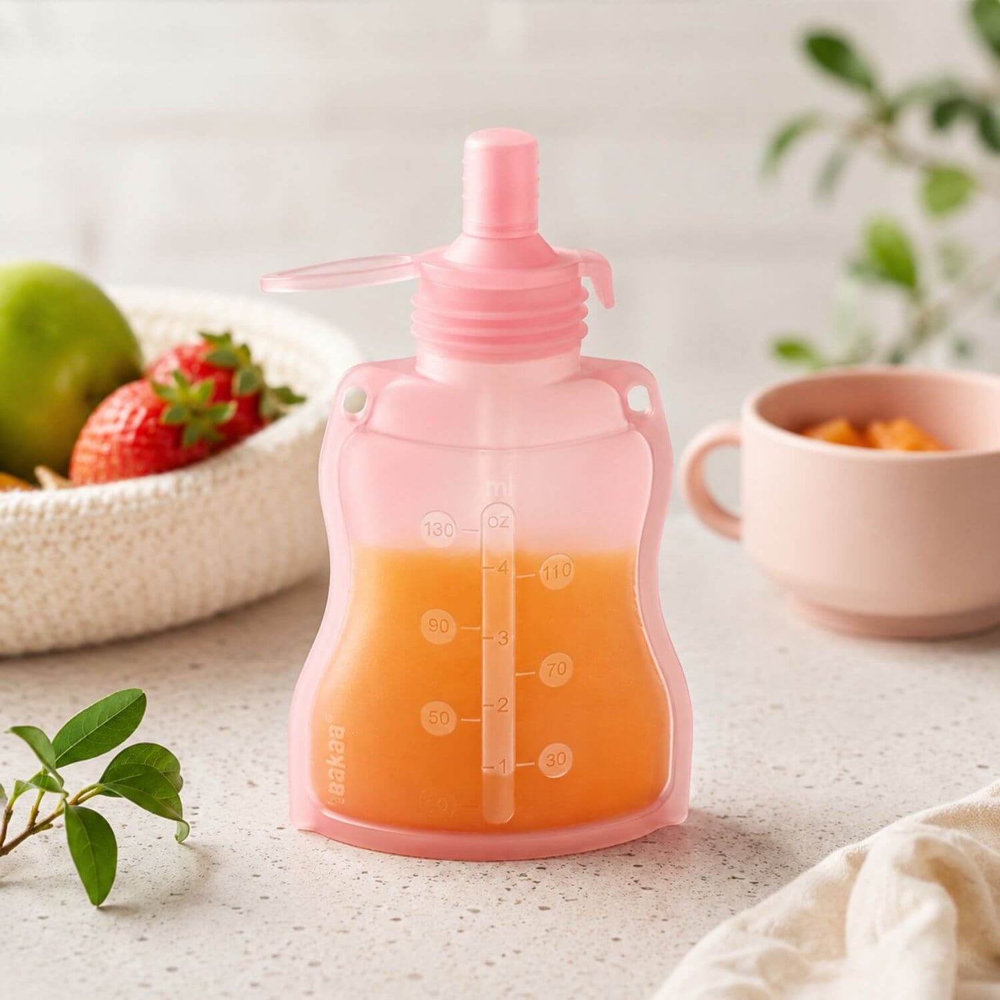 Haakaa Silicone Yummy Pouch with orange juice on a light surface with fruits and a cup in the background.
