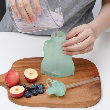 Person pouring liquid into a green silicone pouch on a wooden cutting board with apples and blueberries.
