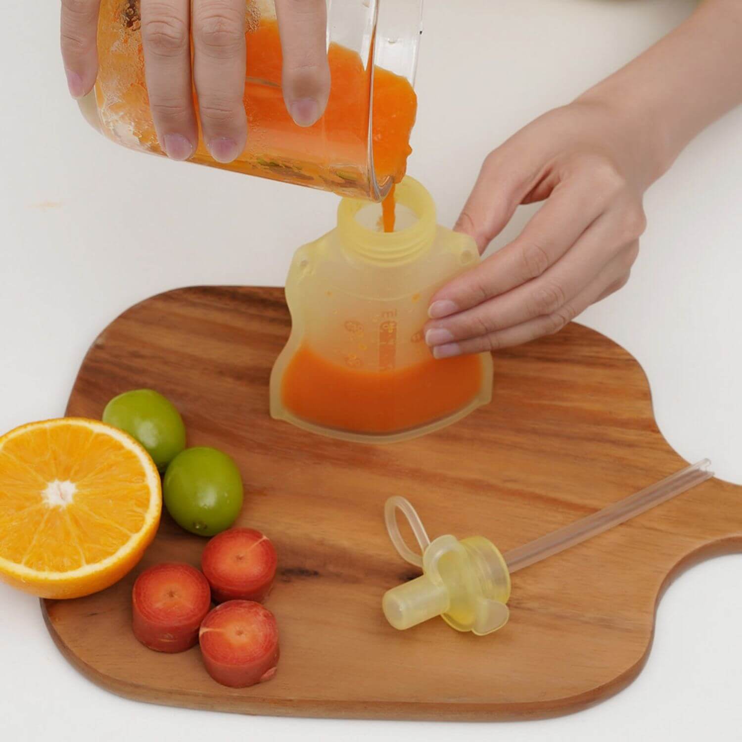 Person pouring orange juice into a yellow silicone pouch on a wooden cutting board with fruits and a pacifier.