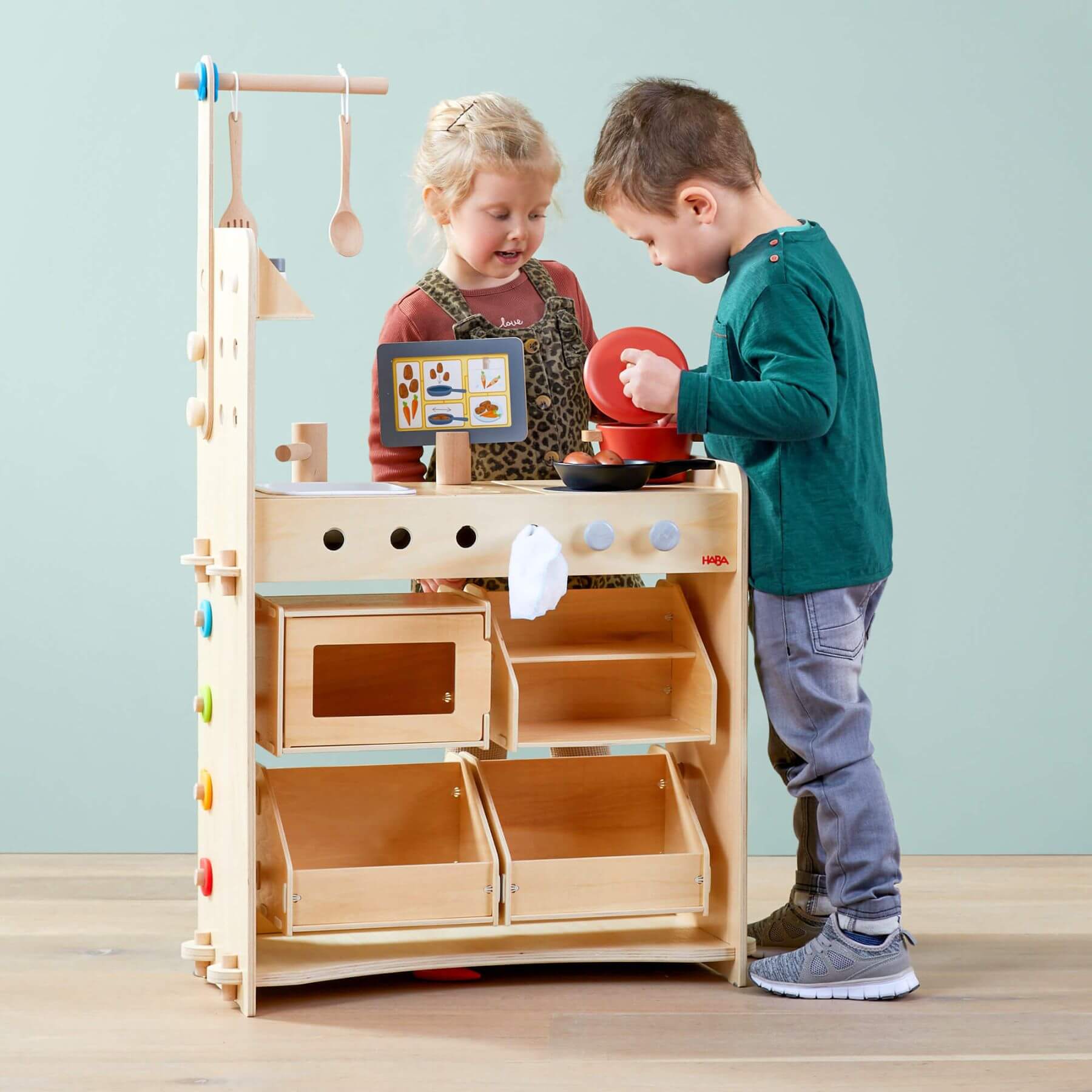 Two children playing with HABA USA Creative Play 3 in 1 – Play Kitchen, Store & Workshop against a light blue wall.