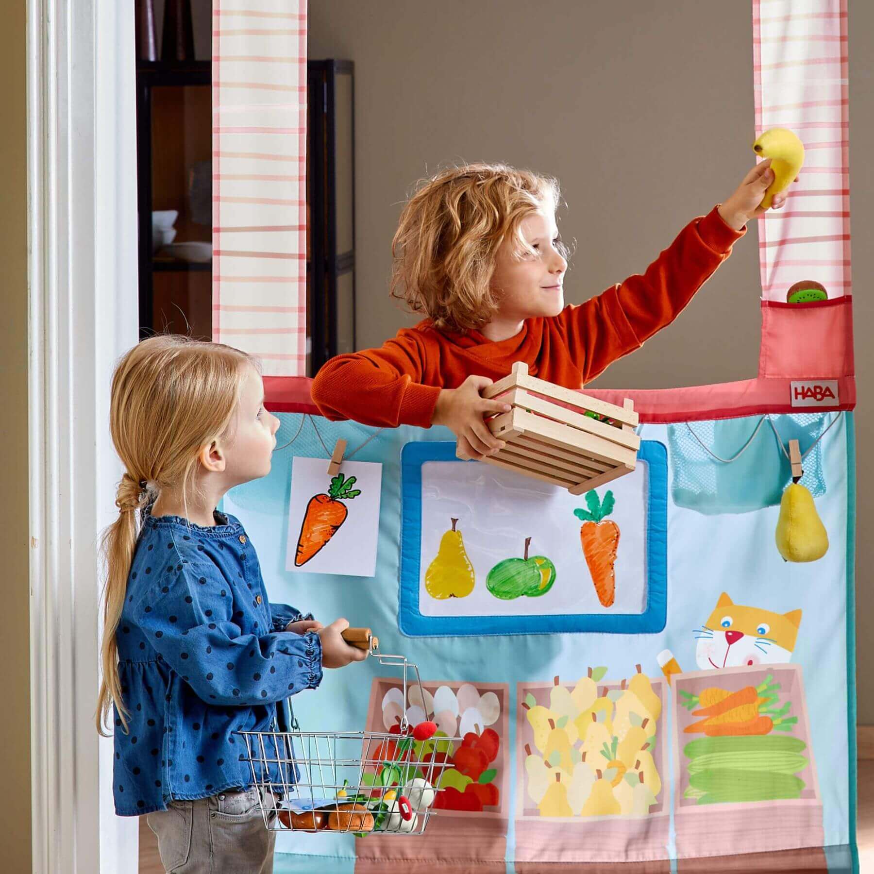 Two children playing with HABA USA Hanging Doorway Play Store, one holding a box and the other with a shopping cart