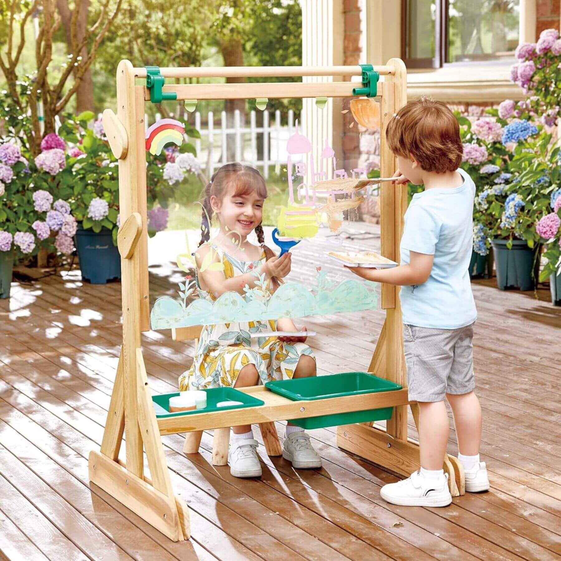 Two children playing with Hape Outdoor Art Easel outdoors on a wooden deck.