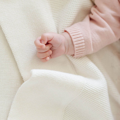 Close-up of a baby's hand in a pink sweater on Saranoni Heirloom Cotton Knit Receiving Blanket Cream