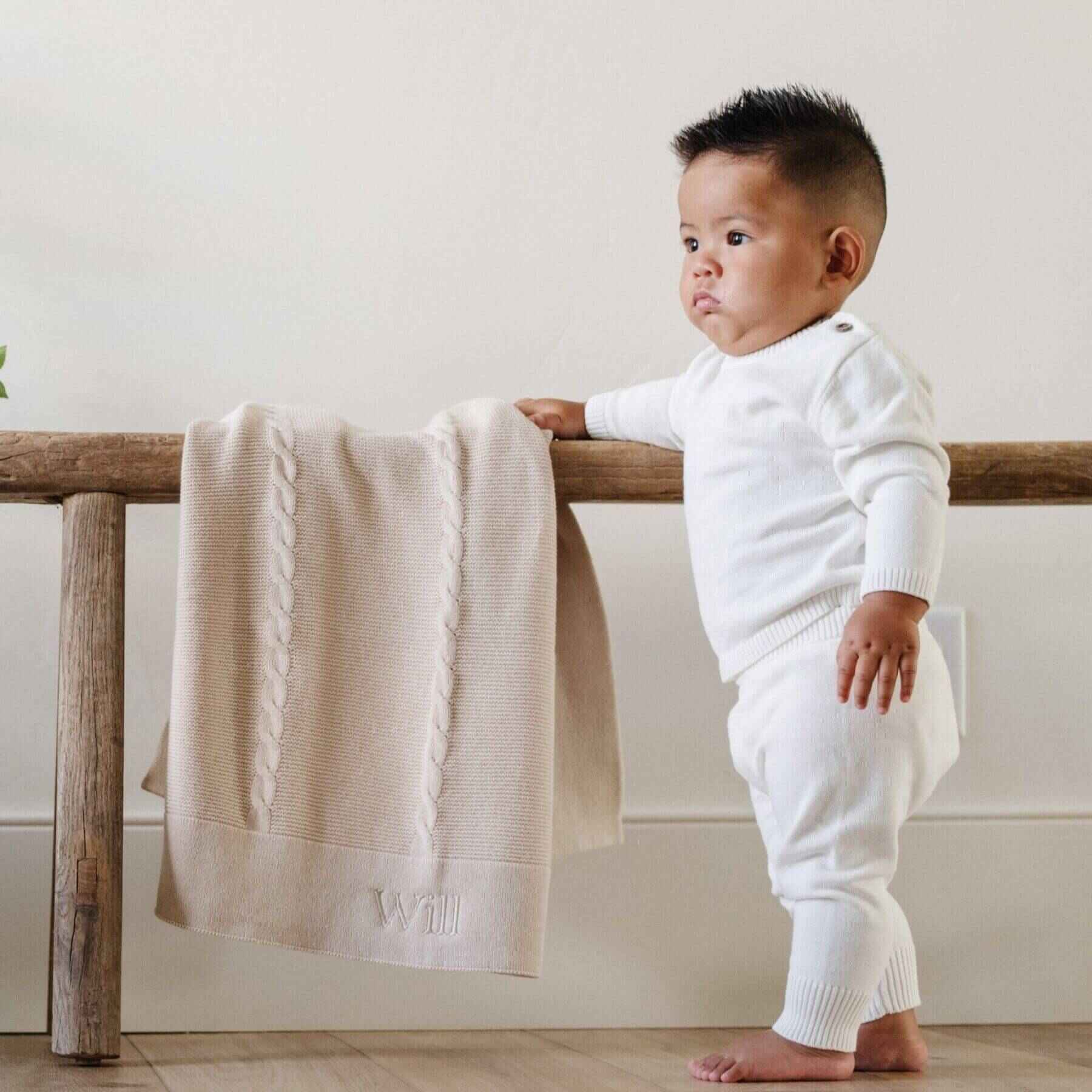 Baby in white outfit leaning against a wooden railing with Saranoni Heirloom Cotton Knit Receiving Blanket