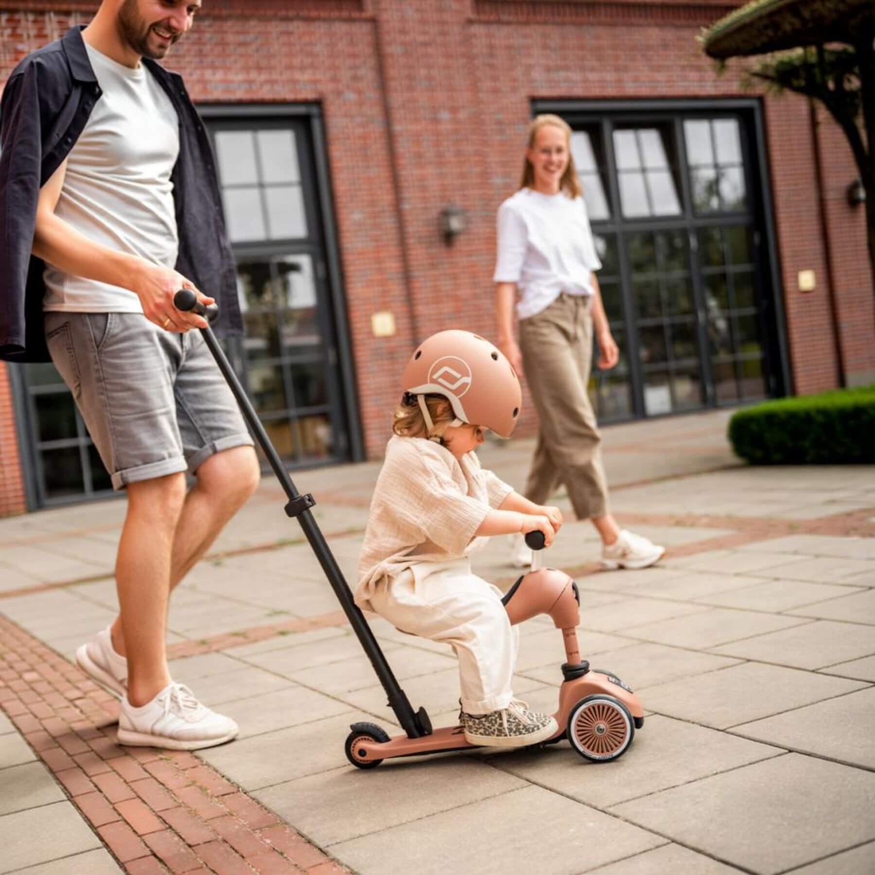 Man pushing a child on a scooter with a pink helmet in an outdoor setting.