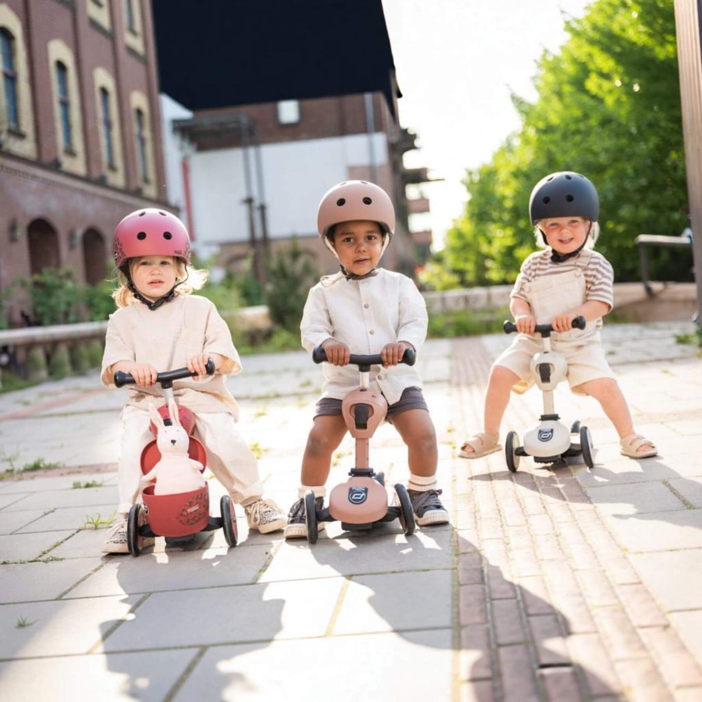 Three children riding scooters on a paved path with buildings and greenery in the background.
