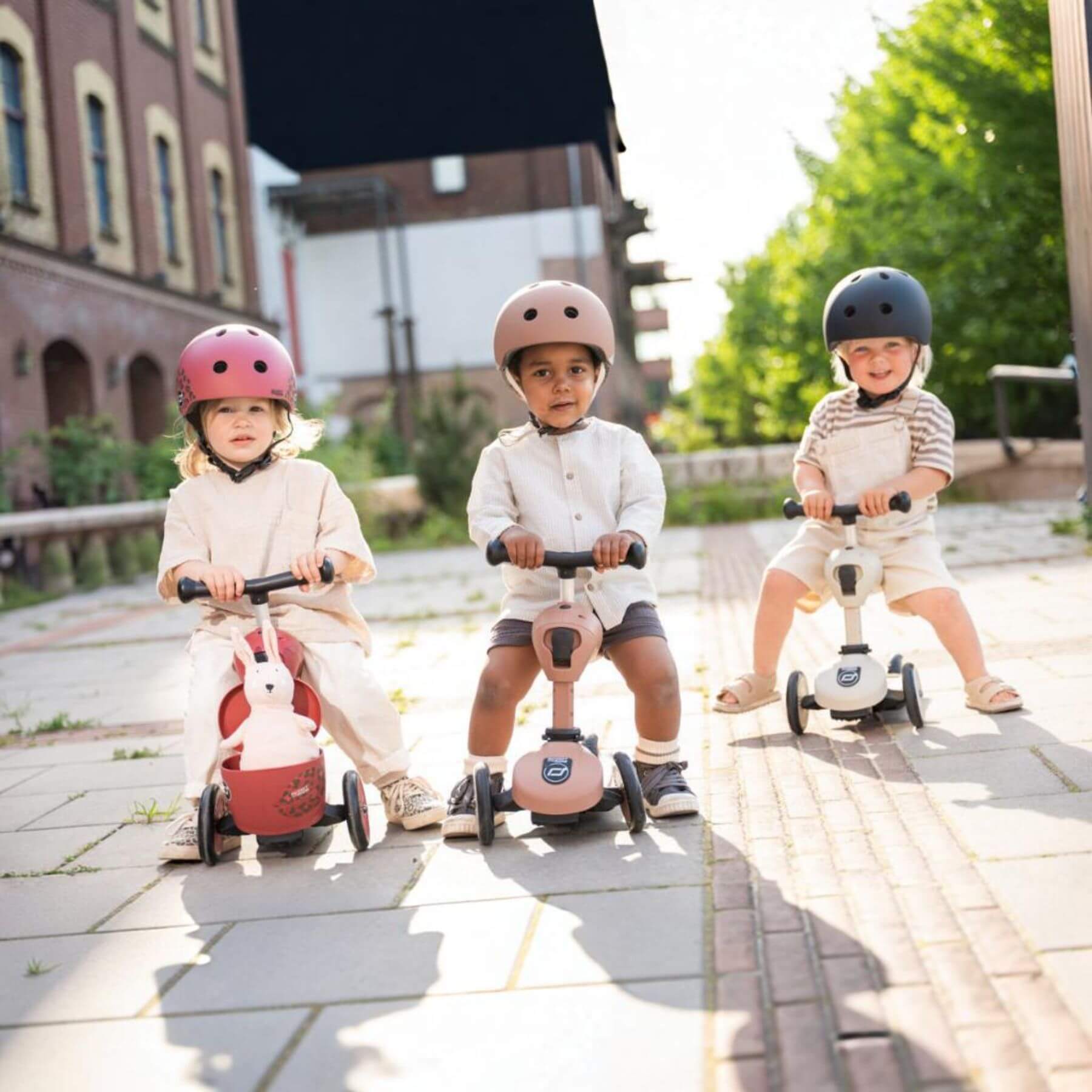 Three children riding scooters on a paved path with buildings and greenery in the background.