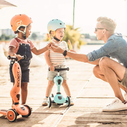 Two children on scooters with a woman on a wooden dock.