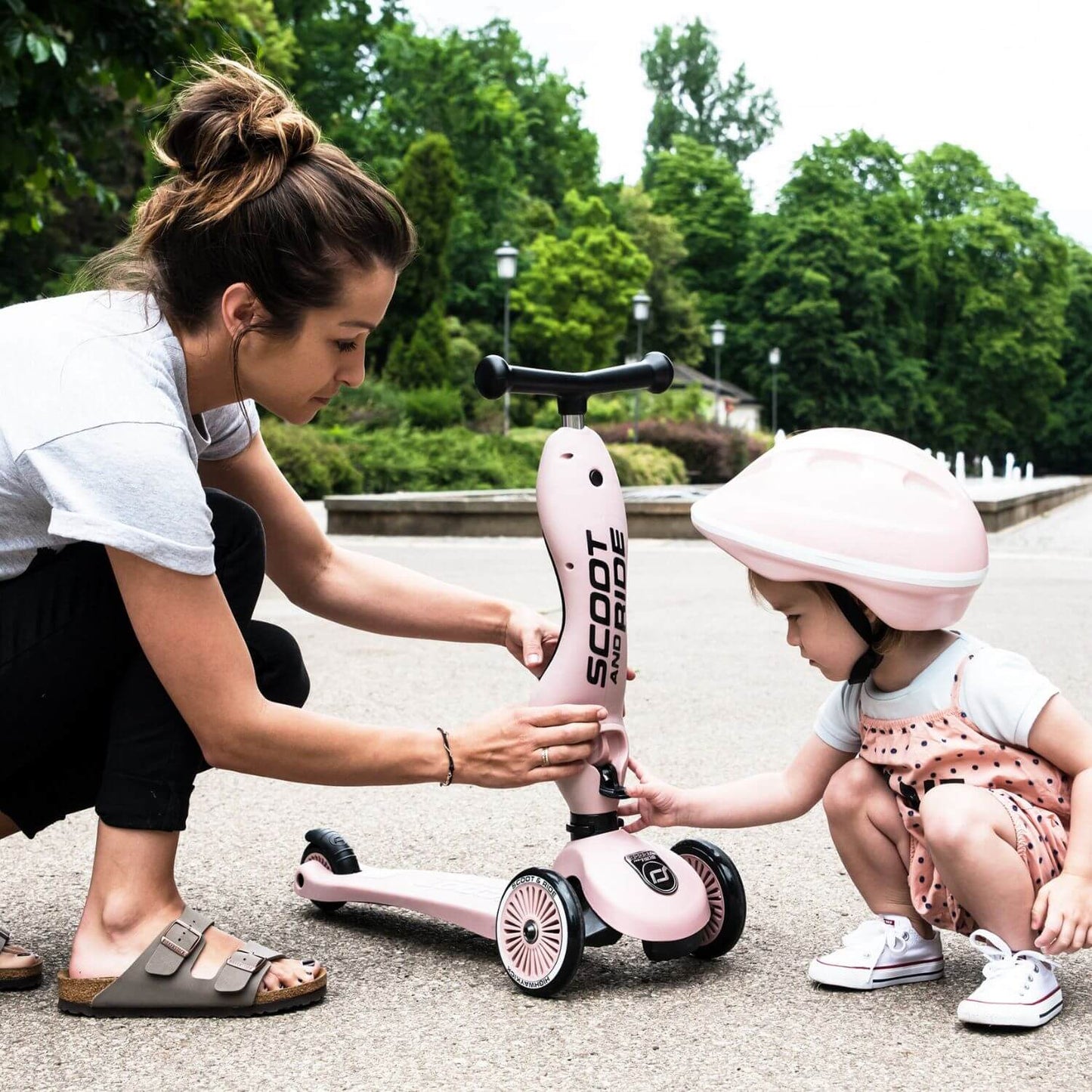 Woman helping a child with a pink scooter in an outdoor setting