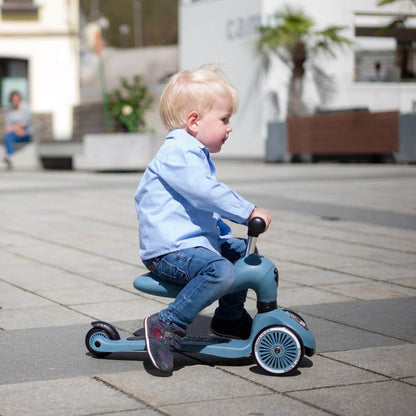 A toddler rides Highwaykick 1 Scooter and Ride On Toy Steel on a paved outdoor plaza.