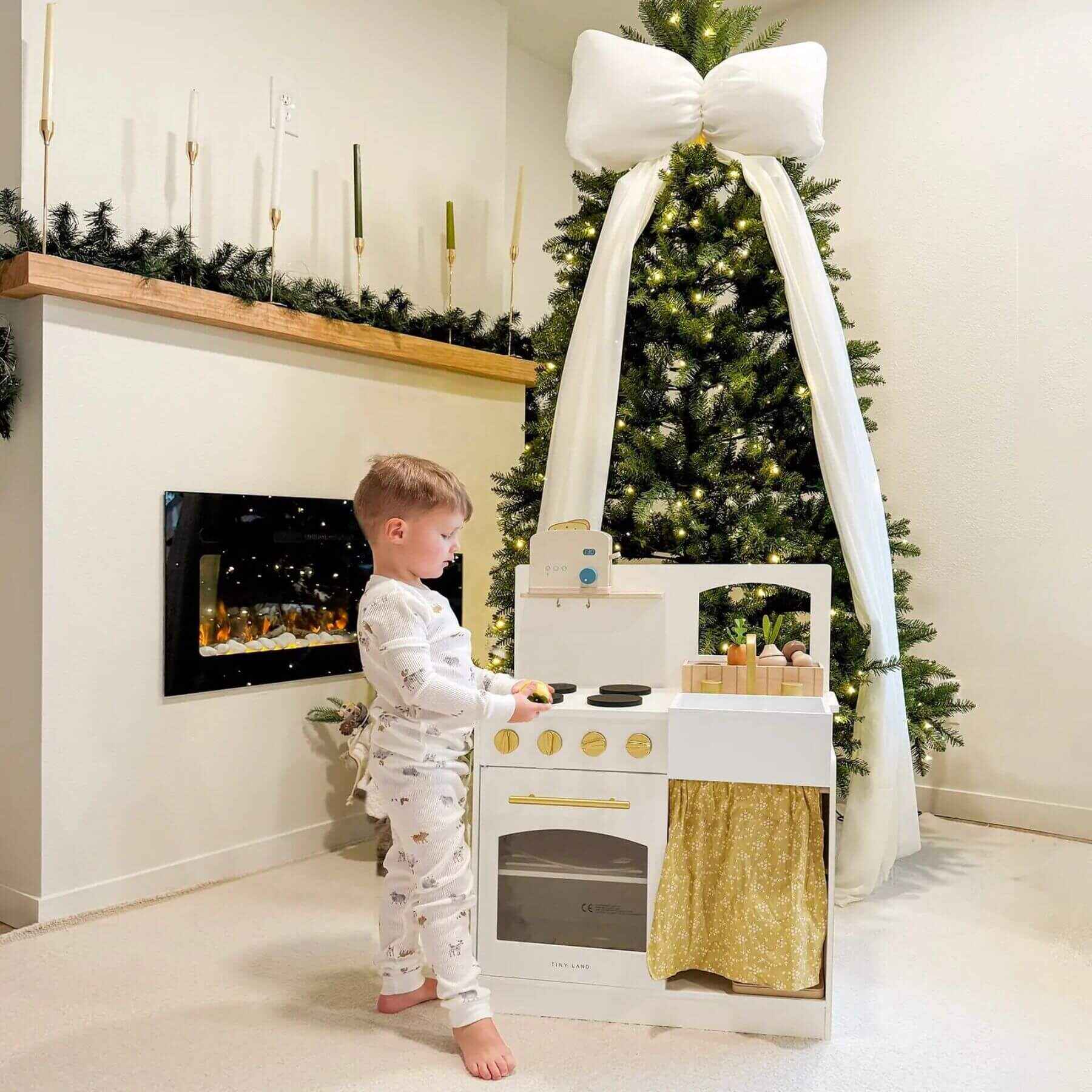Child playing with Tiny Land® Honey Kitchenette - Little Chef's First Kitchen in a room with a Christmas tree and fireplace.