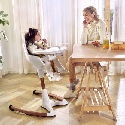 Child in Hot Mom High Chair - White Gold at a dining table with an adult, in a bright room with curtains.