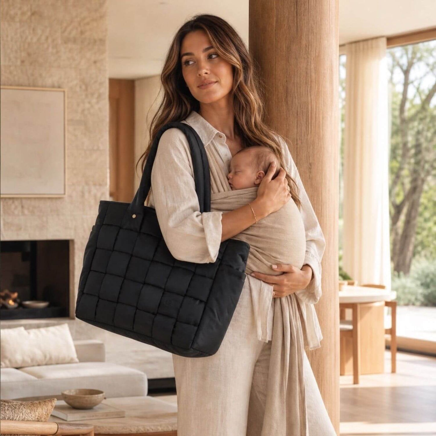 A woman in a neutral-toned outfit carries a baby in a wrap while holding Luli Bebé Infinite Diaper Bag - Black over her shoulder in a bright, modern living room.