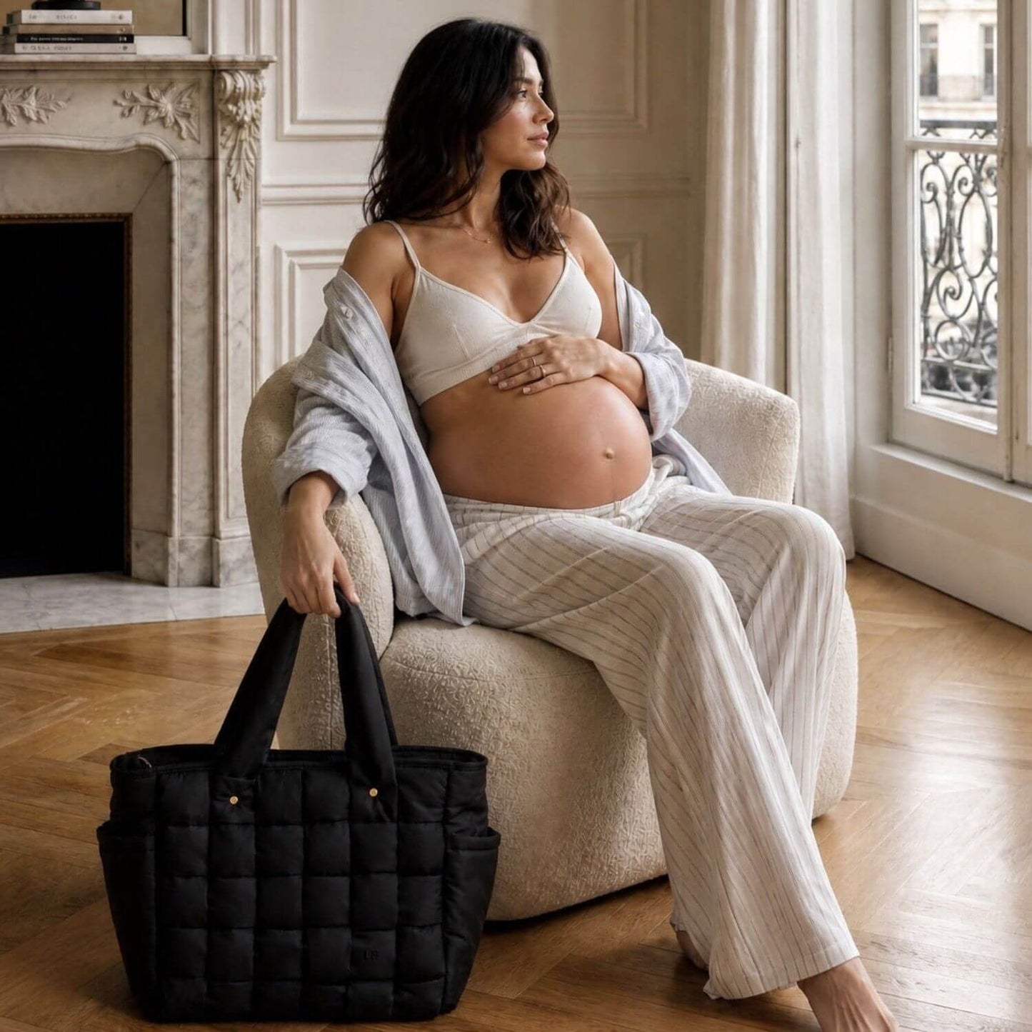 A pregnant woman sits in a cream boucle armchair in a bright, classic Parisian-style room, holding Luli Bebé Infinite Diaper Bag - Black