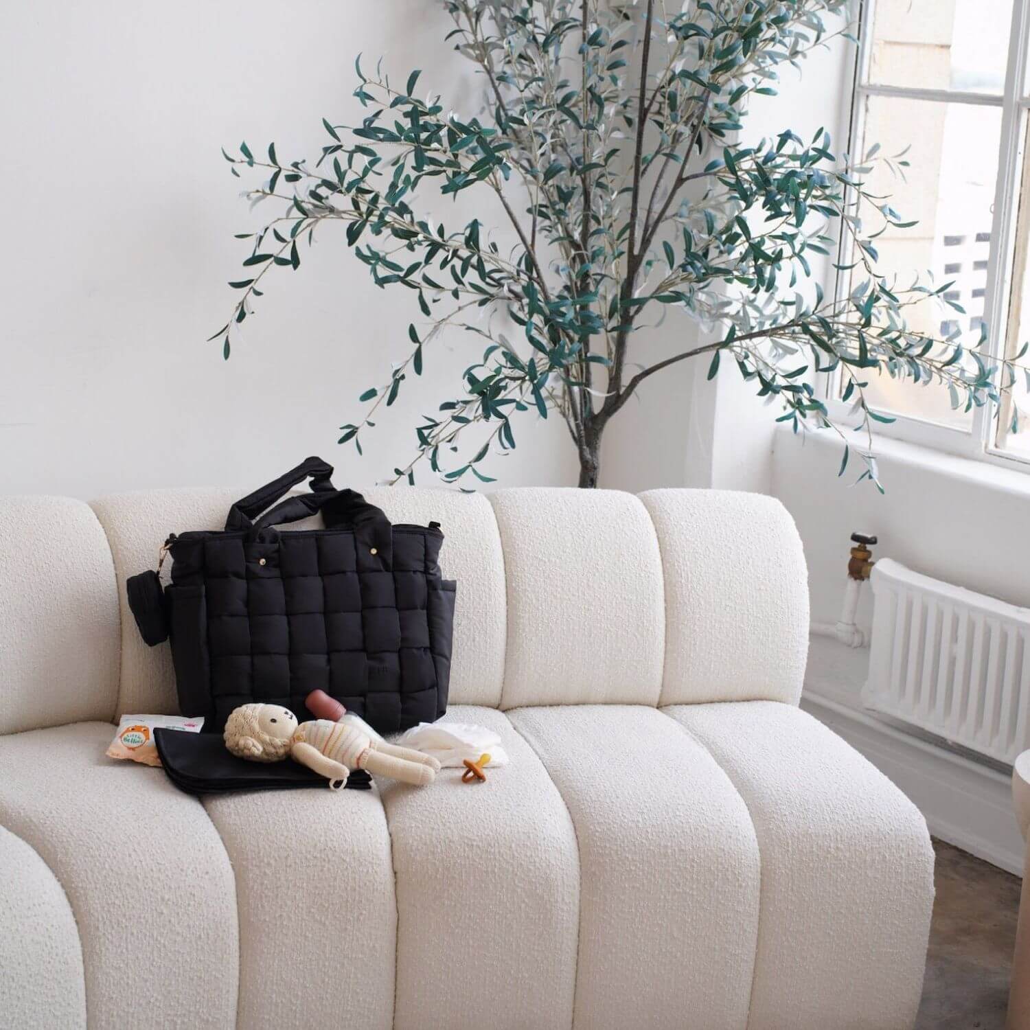Luli Bebé Infinite Diaper Bag - Black sits on a cream boucle sofa in a bright room, styled with a small stuffed doll and a black pouch.