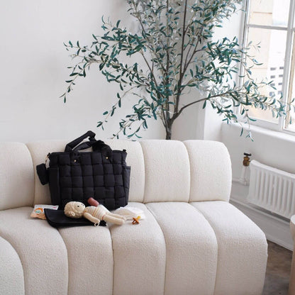 Luli Bebé Infinite Diaper Bag - Black sits on a cream boucle sofa in a bright room, styled with a small stuffed doll and a black pouch.