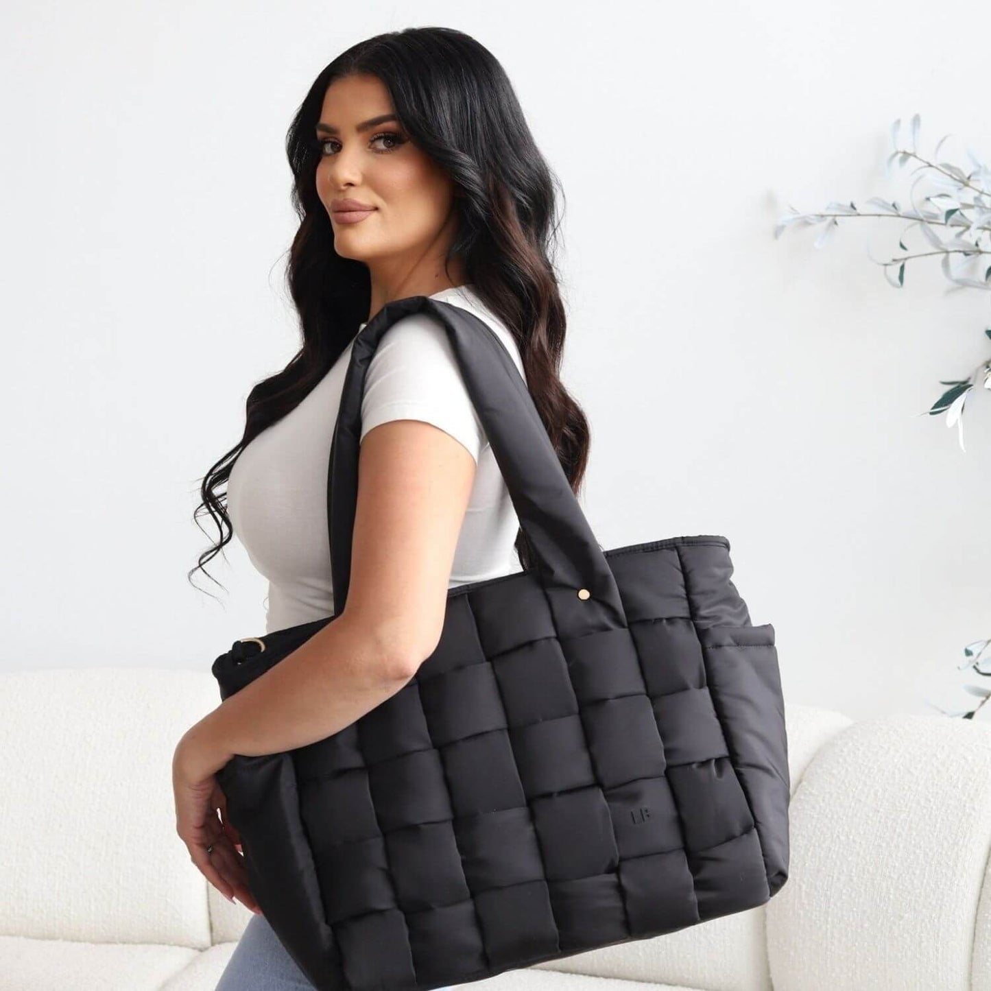 A woman with long dark hair is shown from the side, carrying Luli Bebé Infinite Diaper Bag - Black over her shoulder in a bright, modern room with a white sofa and wall decor.