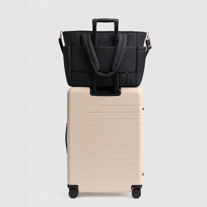 Luli Bebé Infinite Diaper Bag - Black sits on top of a cream-colored hard-shell rolling suitcase against a light gray studio background.