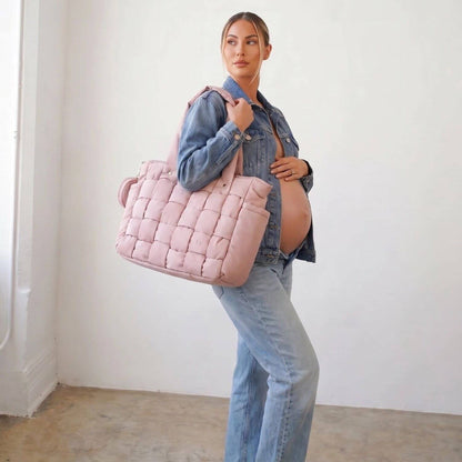 A pregnant woman wearing a denim jacket and jeans carries Luli Bebé Infinite Diaper Bag - Blush against a plain white wall.