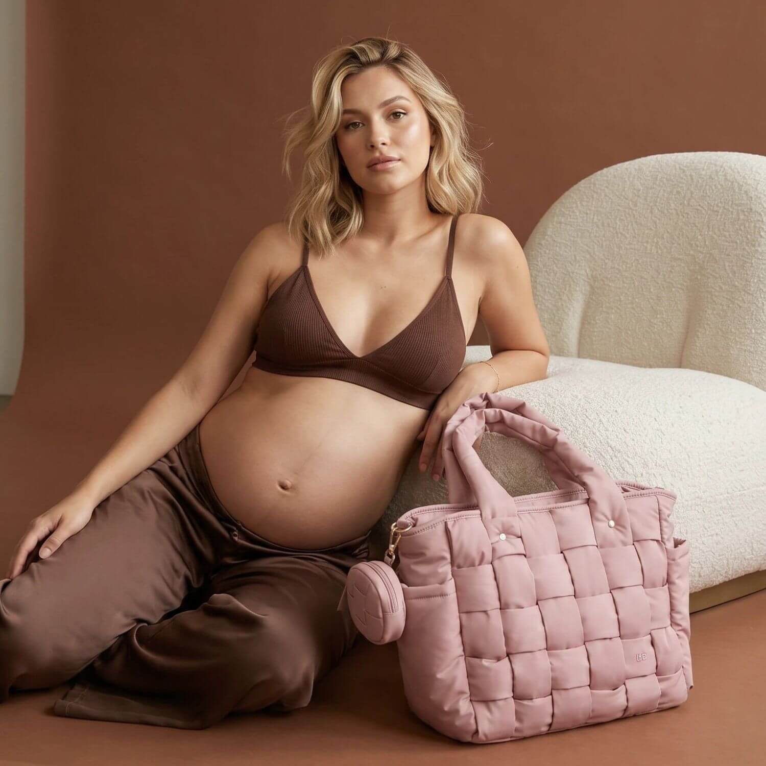 A pregnant woman in a brown bra and matching pants sits on a cream boucle chair next to Luli Bebé Infinite Diaper Bag - Blush