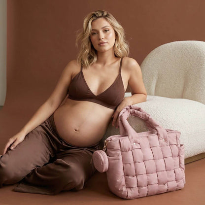 A pregnant woman in a brown bra and matching pants sits on a cream boucle chair next to Luli Bebé Infinite Diaper Bag - Blush