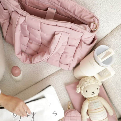 Luli Bebé Infinite Diaper Bag - Blush is shown in a styled flat-lay arrangement on a light beige carpet, surrounded by a cream travel mug, a small pink bottle, a plush sheep toy, and a white bag held by a hand.