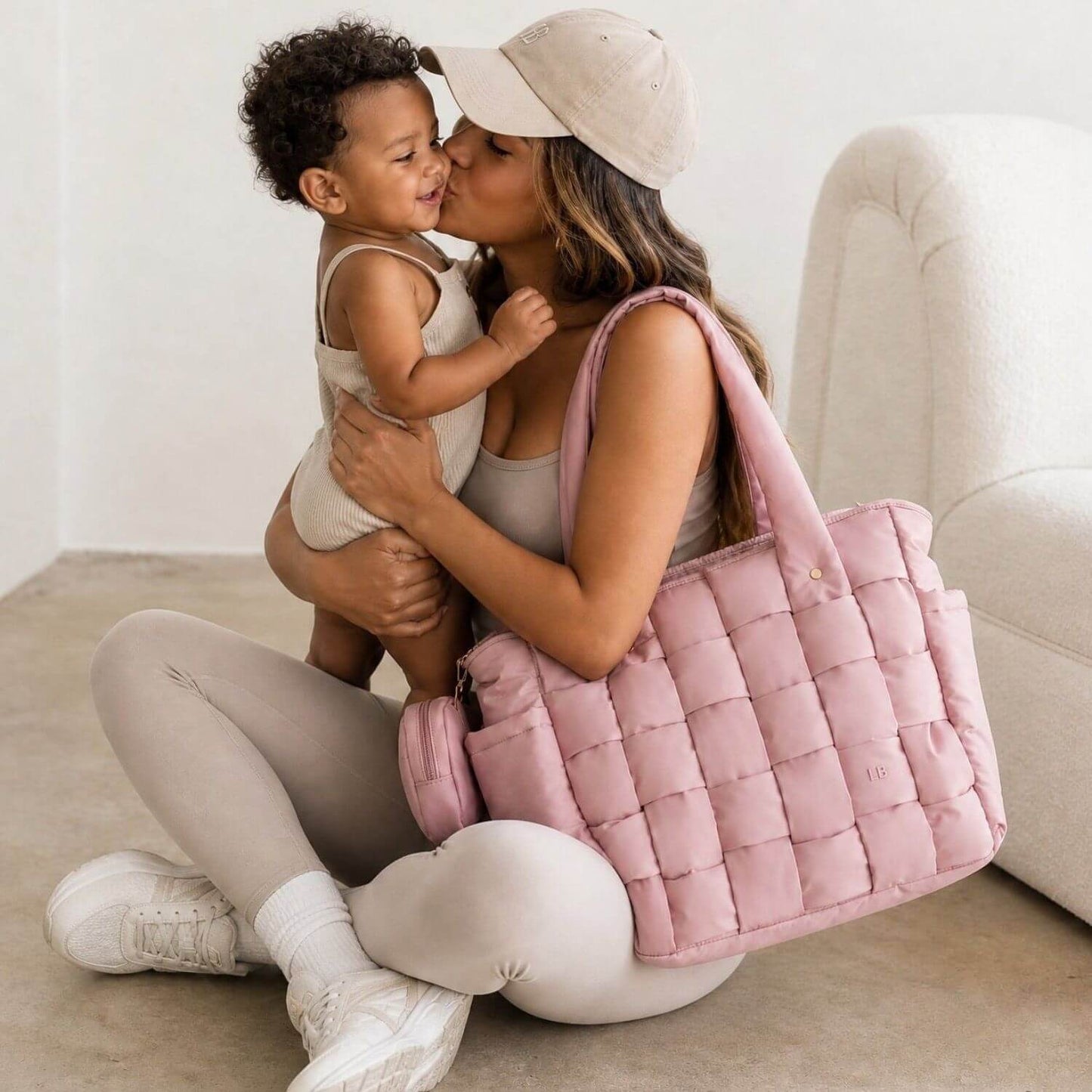 A woman in a beige cap and matching loungewear holds a baby while carrying Luli Bebé Infinite Diaper Bag - Blush