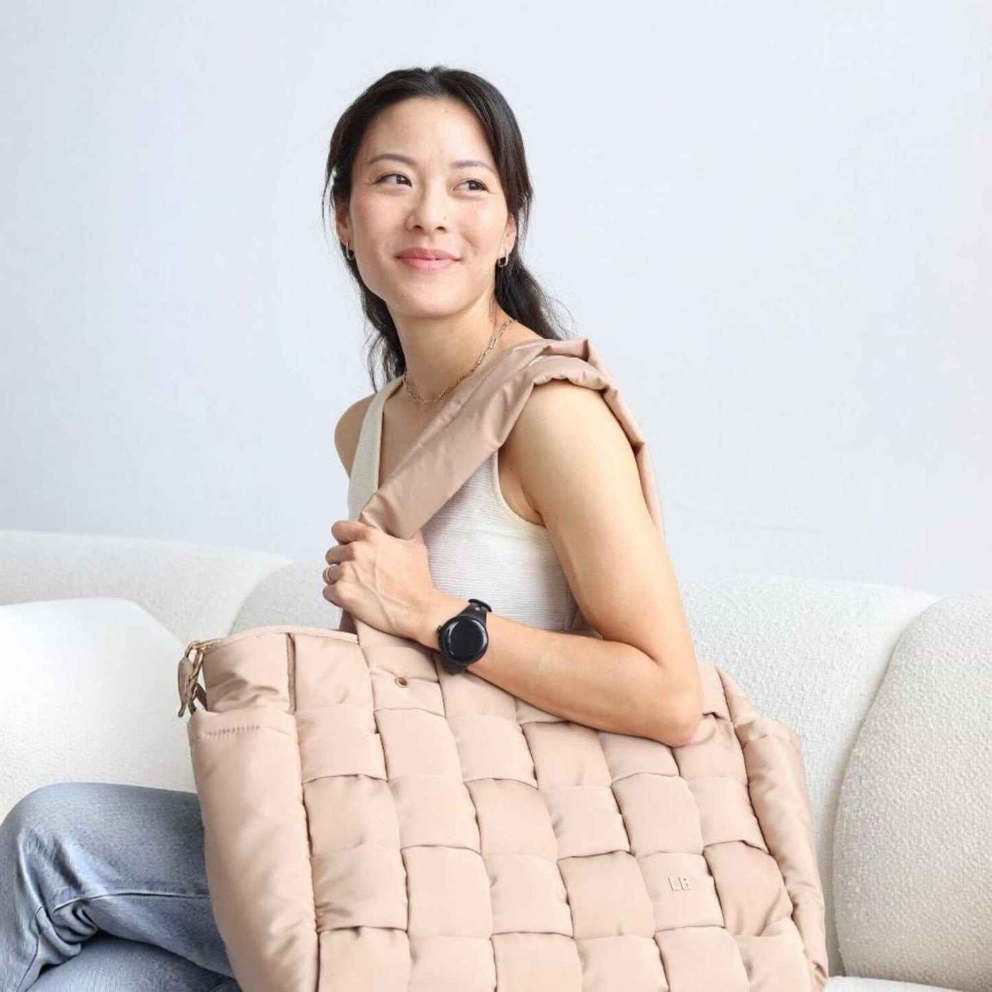 A woman sitting on a white sofa holds Luli Bebé Infinite Diaper Bag - Latte
