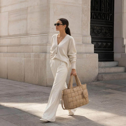 A woman in a cream knit cardigan and matching wide-leg trousers walks outdoors carrying Luli Bebé Infinite Diaper Bag - Latte