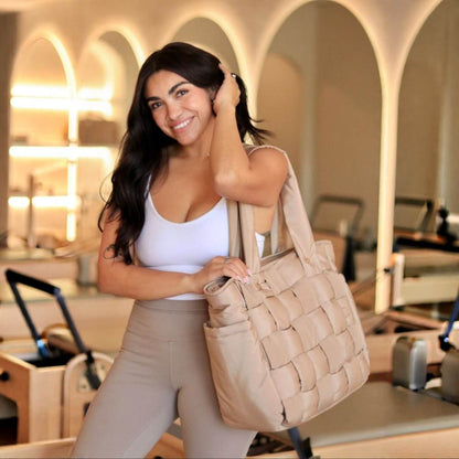 A woman in a white tank top and beige leggings holds Luli Bebé Infinite Diaper Bag - Latte in a bright, modern studio with arched windows in the background.
