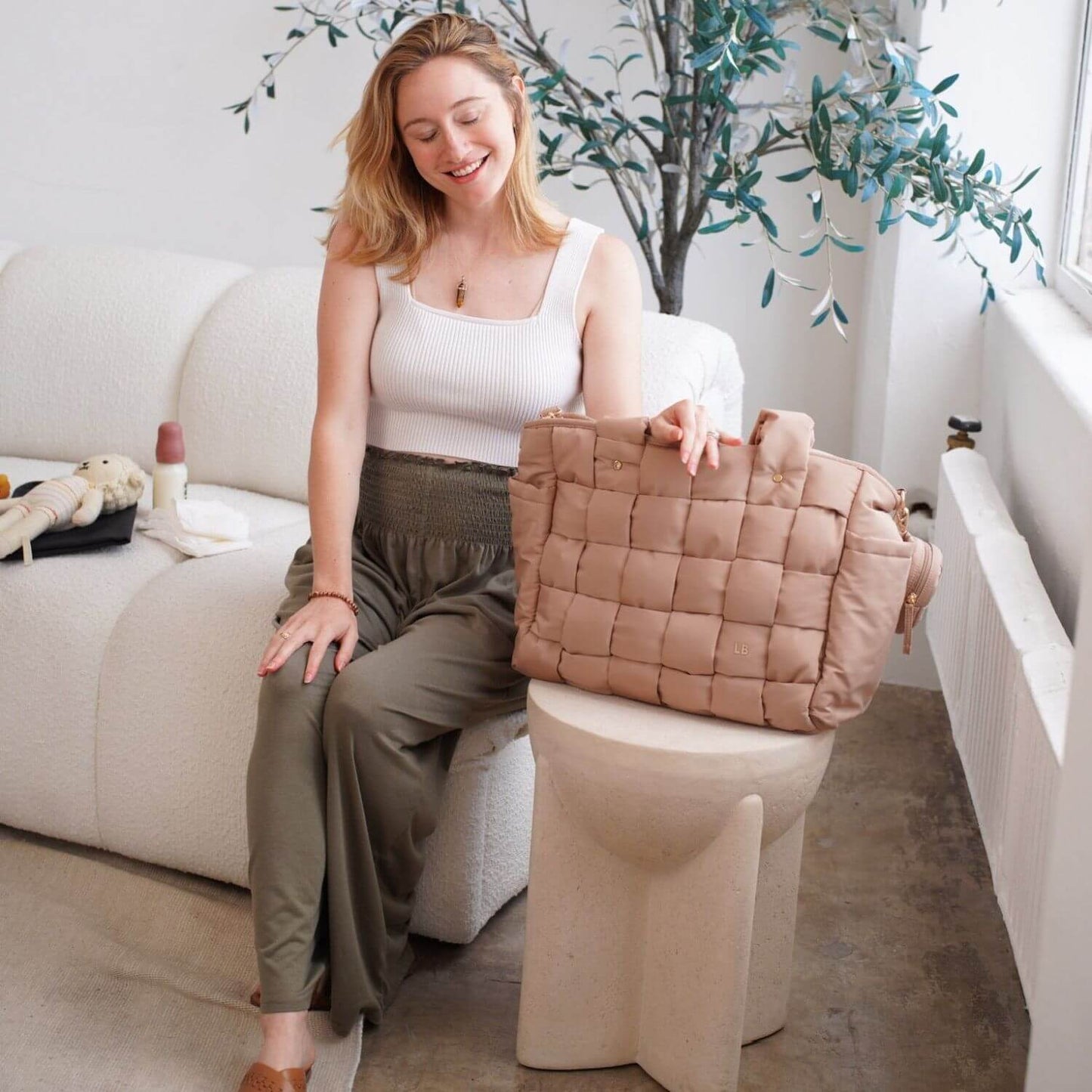 A woman sits on a cream boucle sofa in a bright room, holding Luli Bebé Infinite Diaper Bag - Latte resting on a sculptural side table.