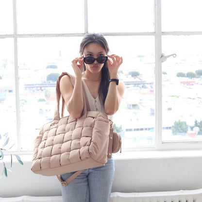 A woman wearing black sunglasses and a white top stands in front of a bright window, carrying Luli Bebé Infinite Diaper Bag - Latte