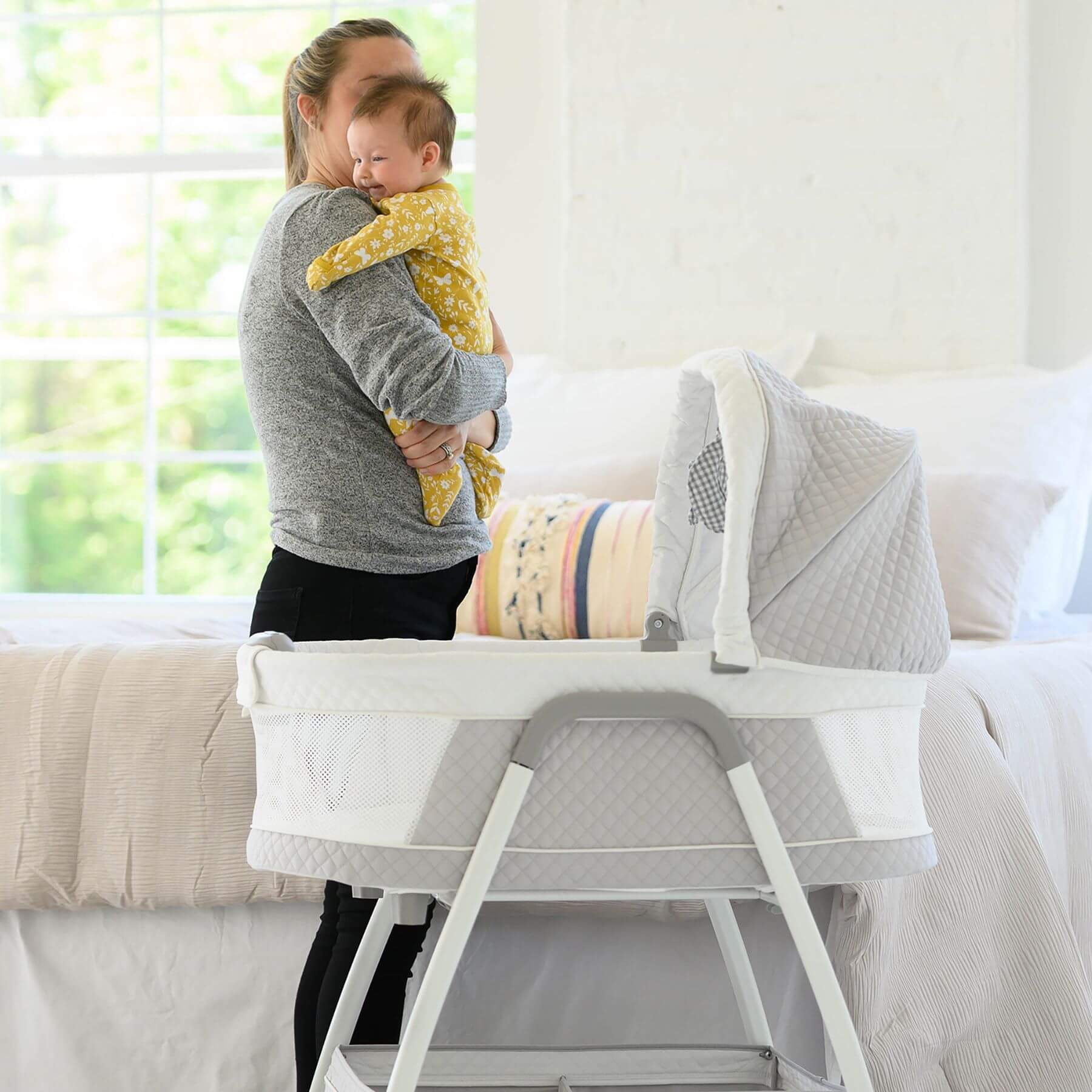 Mom Holding Baby next to TruBliss Journey 3-in-1 Bassinet