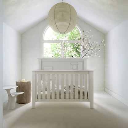 "Just White" Geometric Convertible Crib in a bright room with a large window and decorative lamp.