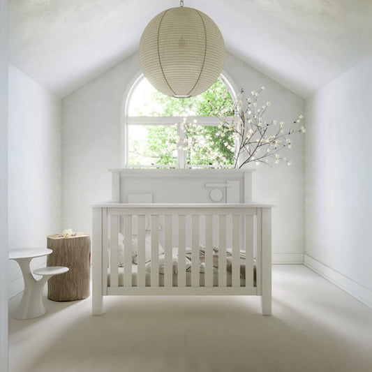 "Just White" Geometric Convertible Crib in a bright room with a large window and decorative lamp.