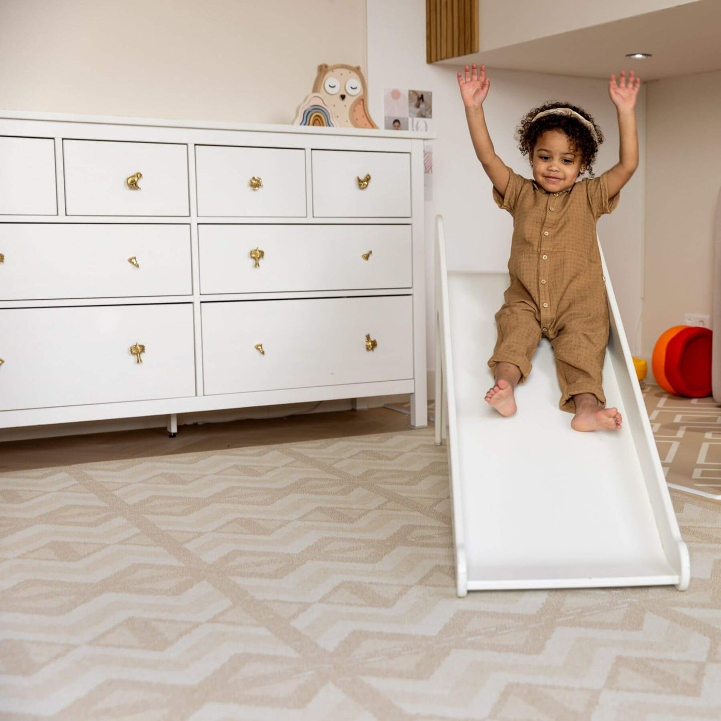 Child sliding down a white plastic slide in a room with a white dresser and colorful toys.