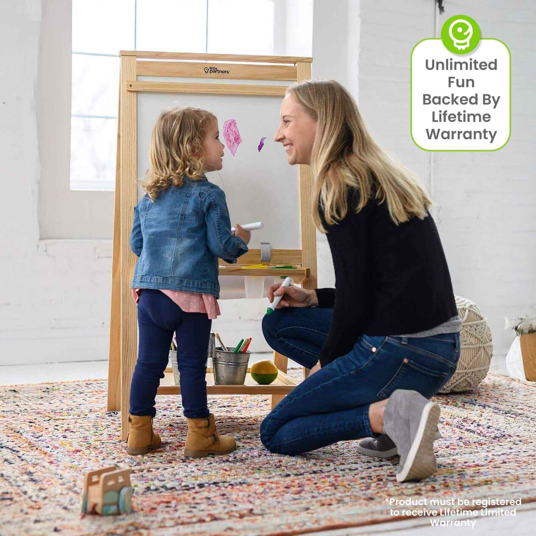 Woman and child playing with a Little Partners Folding Artist EZ Easel Natural in a room with a colorful rug.