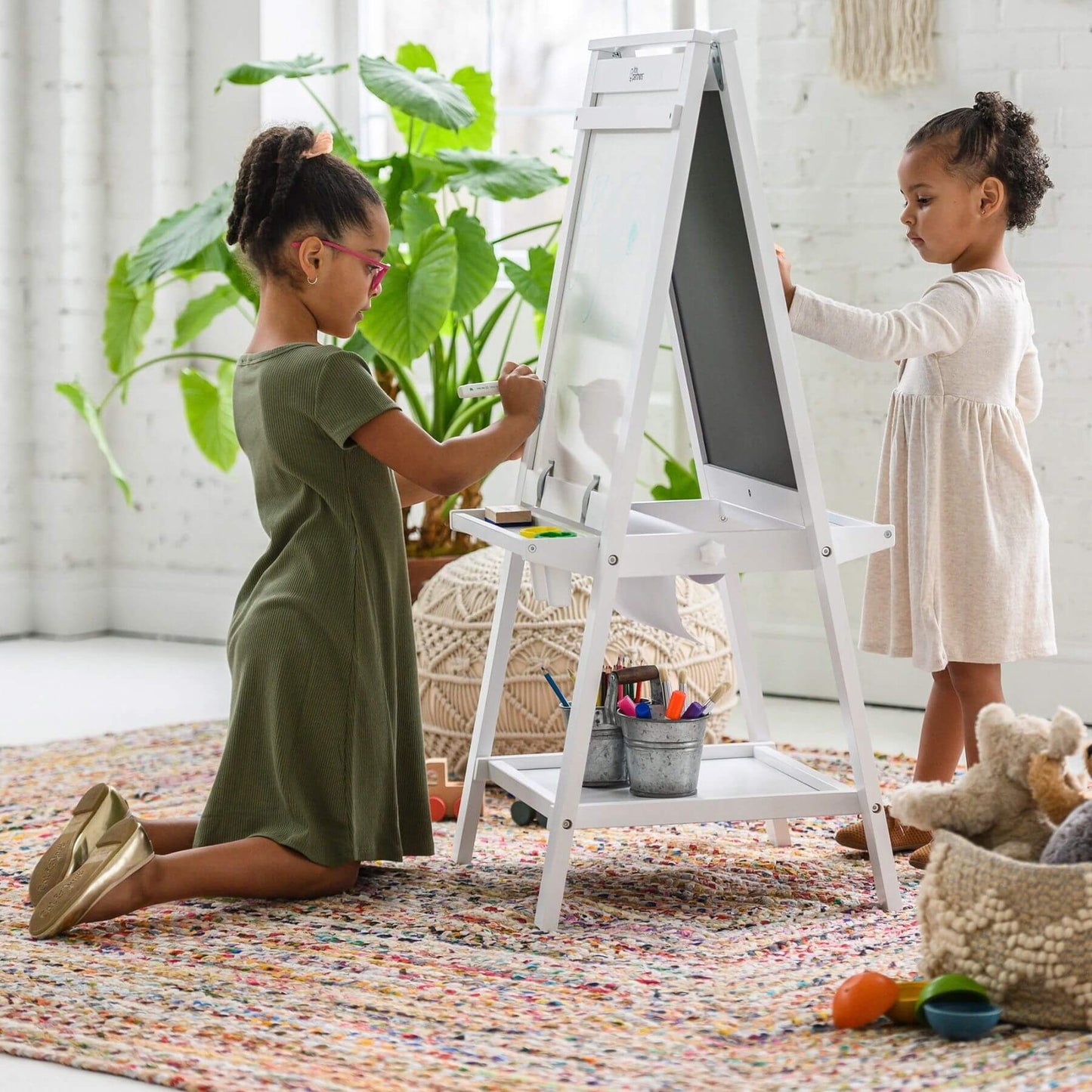 Two young girls painting on a Little Partners Folding Artist EZ Easel White in a bright room with plants and toys.