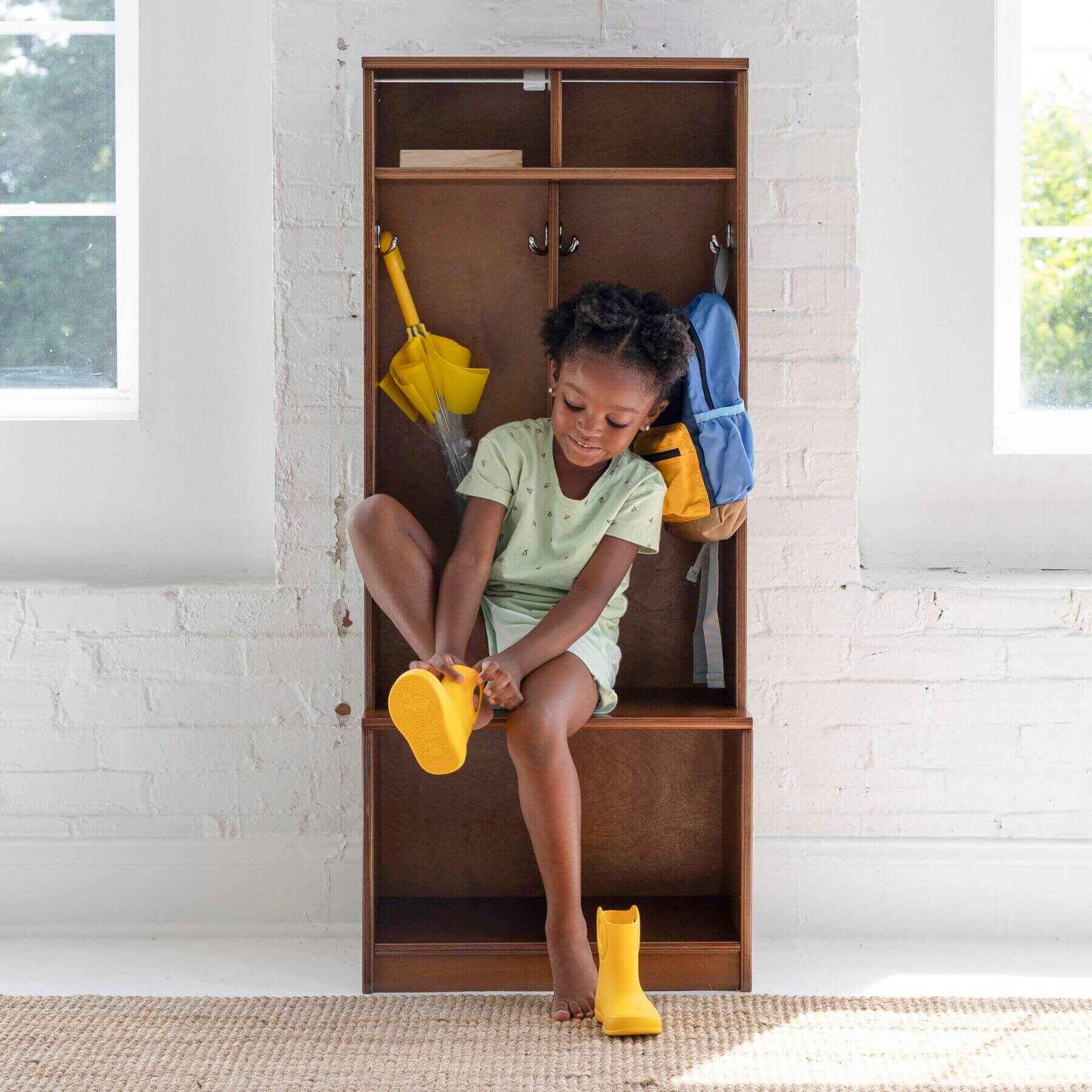 Child sitting on Little Partners Learn 'N Store Cubby with shoes and backpacks, in a room with white brick walls.