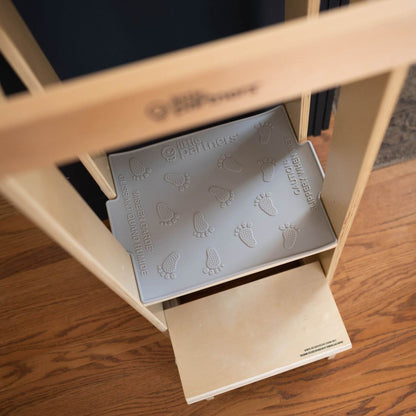 Wooden shelf with a Little Partners Silicone Mat featuring footprints and text, on a wooden floor.