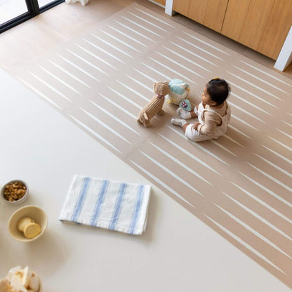 A baby sits on Eeveve Lucio Play Mat - Desert Sand in a bright room, playing with plush toys.