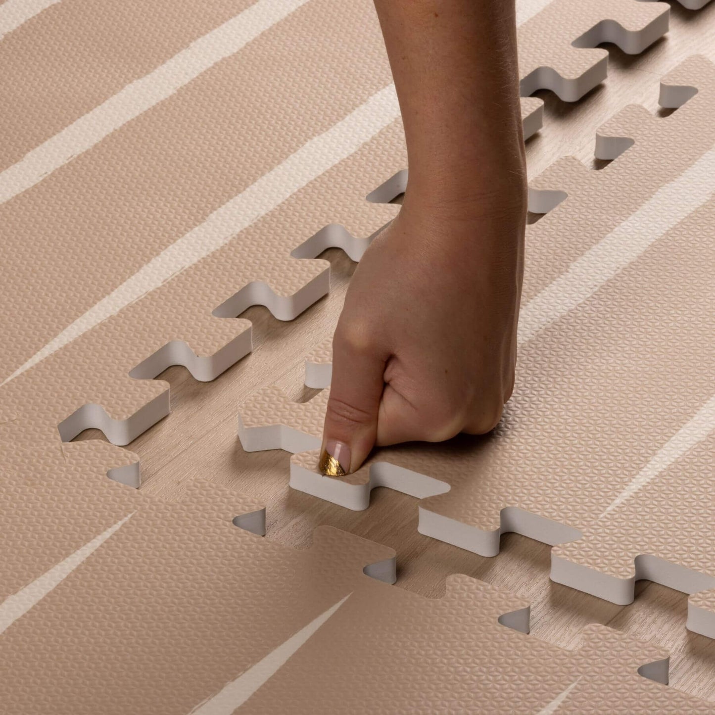A close-up of a person's hand pressing down on interlocking textured foam floor tiles in a beige and off-white pattern.
