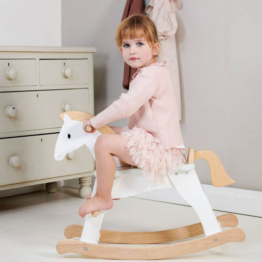 Young girl sitting on Tender Leaf Lucky Rocking Horse in a room with a dresser.