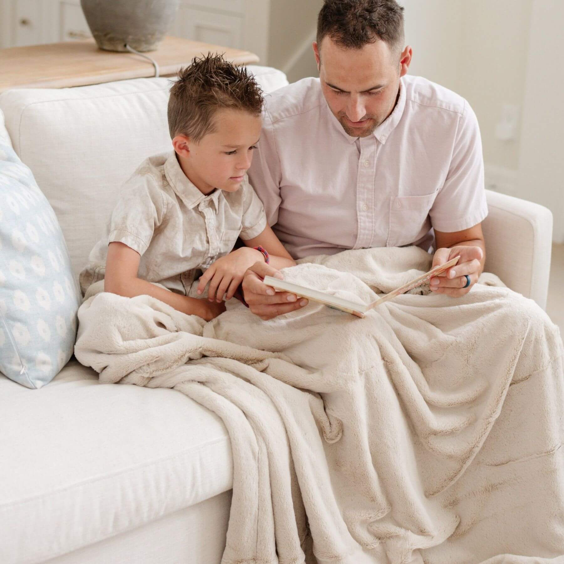 Man and child sitting on a couch together, reading a book, with Saranoni Lush Throw Blanket draped over them.