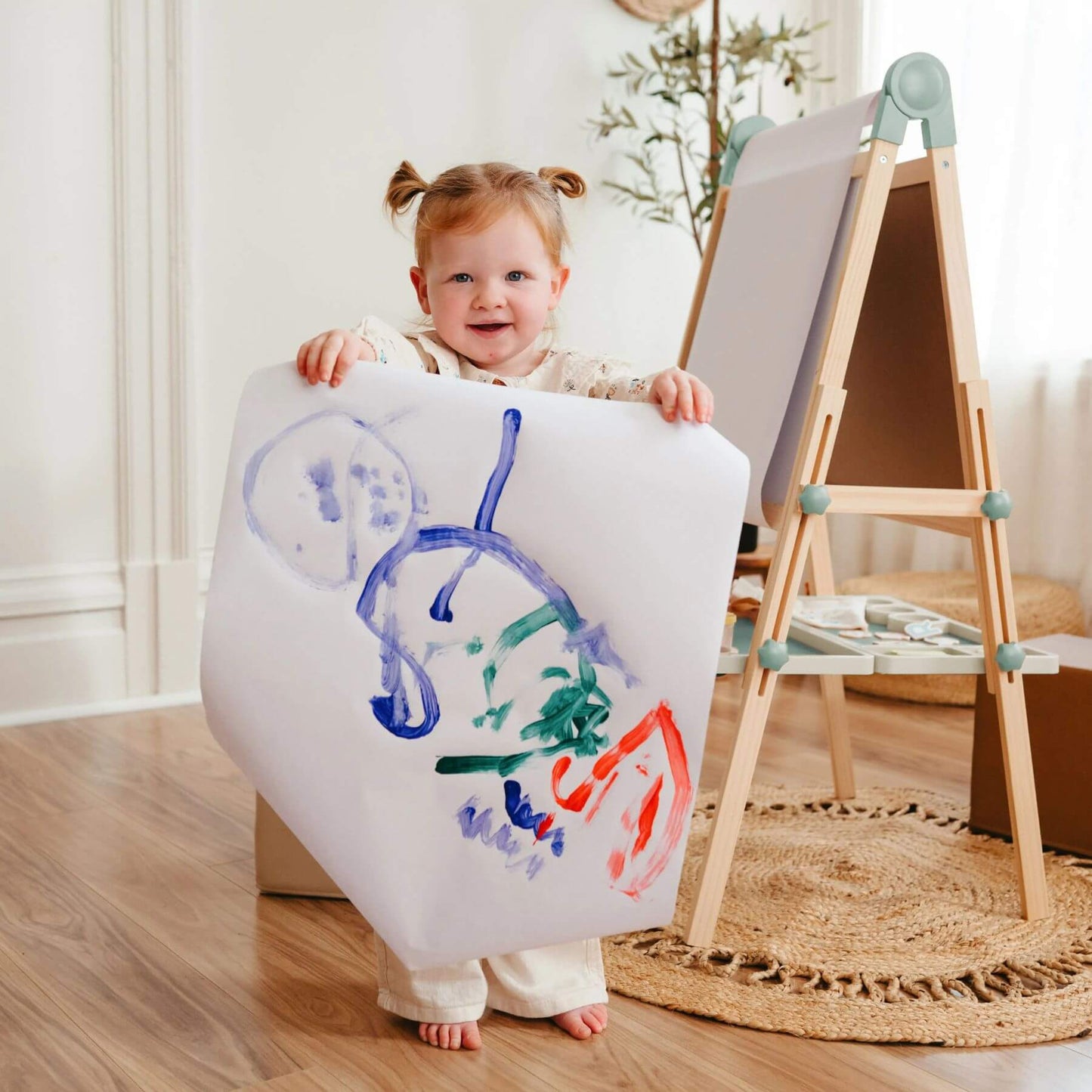 Child holding a large piece of paper with colorful drawings in a room with a wooden floor and a plant.