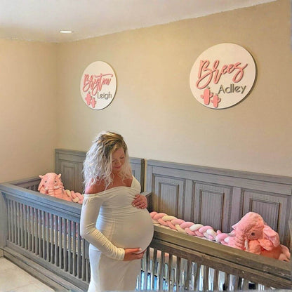 Pregnant woman standing in a nursery with two cribs and decorative signs on the wall.