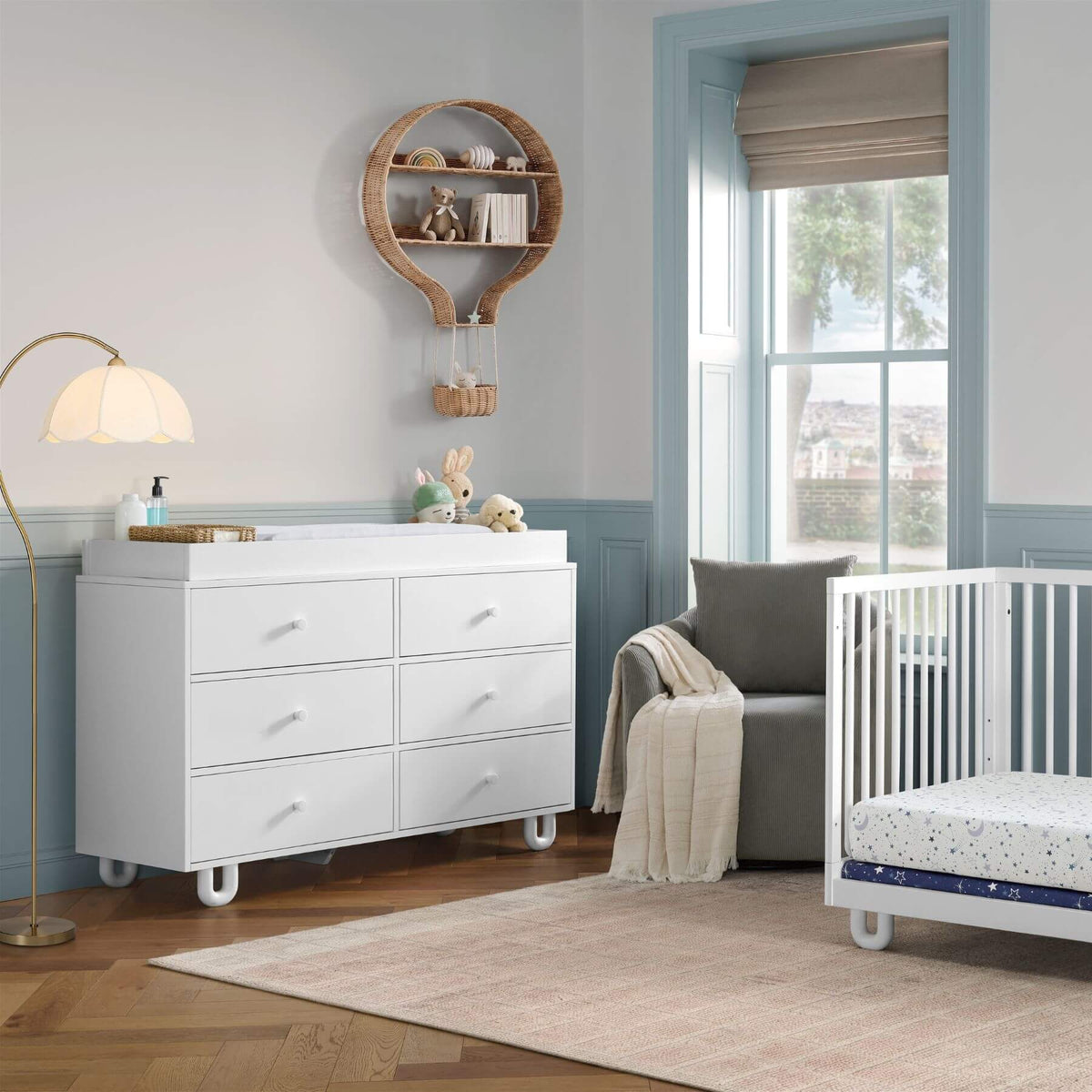 Soho Baby Meridian 6 Drawer Dresser - White, styled in a nursery next to a white crib, a gray armchair, a floor lamp, and a window with blue trim.