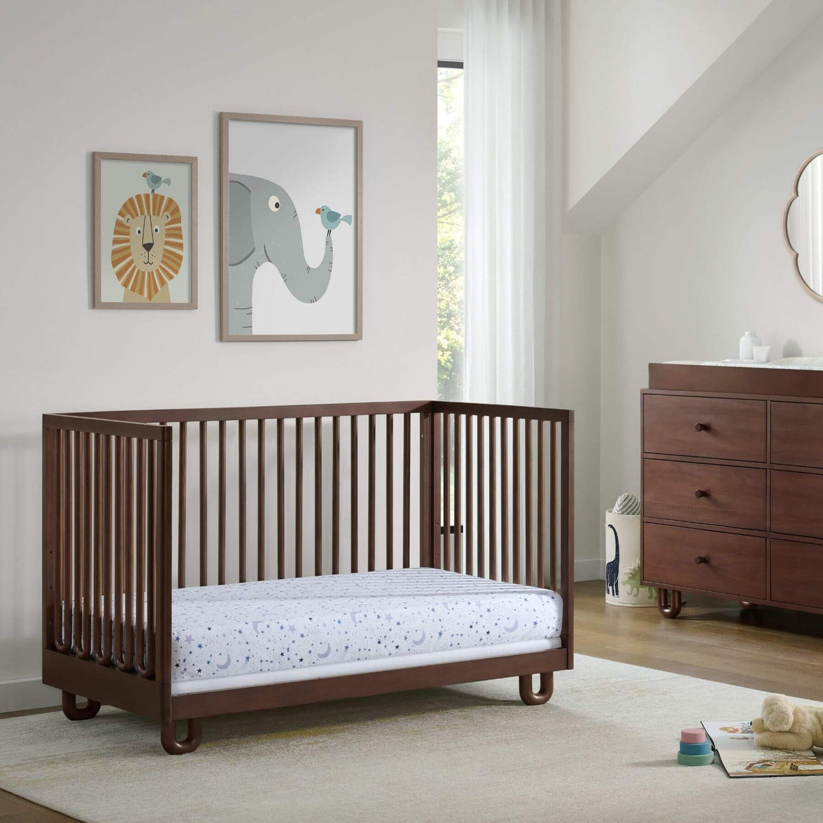 Soho Baby Meridian Island Crib - Cognac with a mattress, shown in a styled nursery setting with a dresser, framed animal art, and a window.