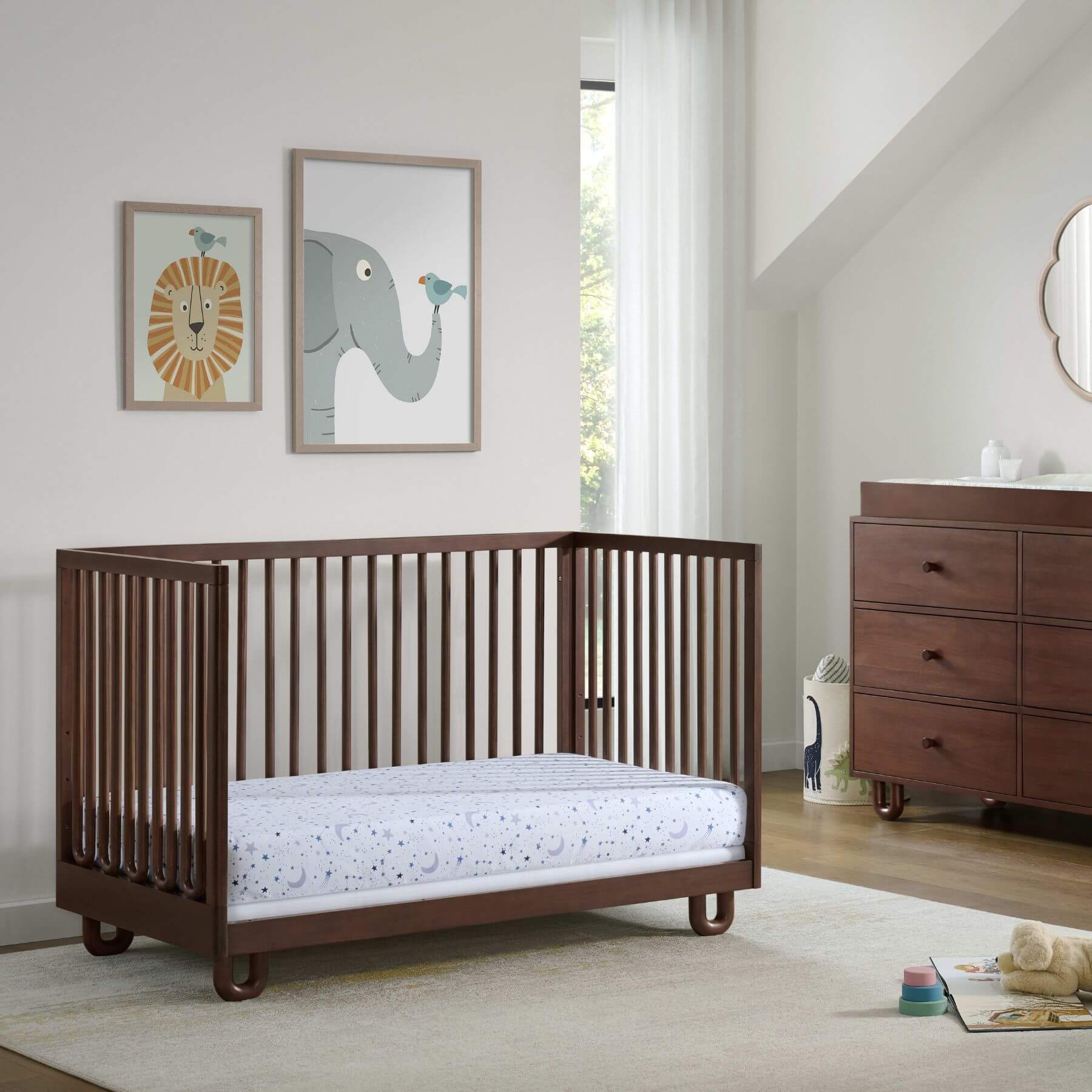 Soho Baby Meridian Island Crib - Cognac with a mattress, shown in a styled nursery setting with a dresser, framed animal art, and a window.